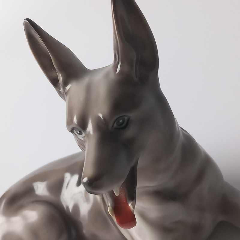 figurka porcelanowa owczarek niemiecki Zbliżenie na głowę porcelanowego psa, widoczny czerwony język i malowane oczy.