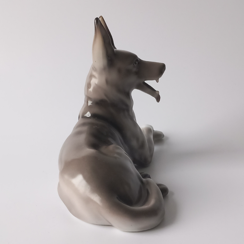 figurka porcelanowa owczarek niemiecki Bok leżącego wilczura z porcelany, szare umaszczenie, otwarty pysk.