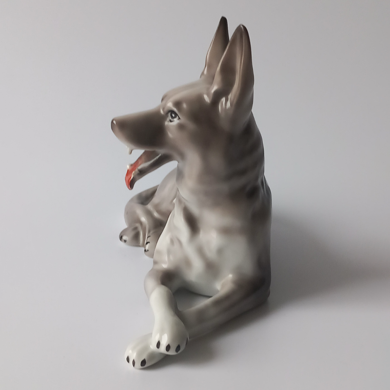 figurka porcelanowa owczarek niemiecki Profil leżącego wilczura z porcelany, szare umaszczenie, otwarty pysk.