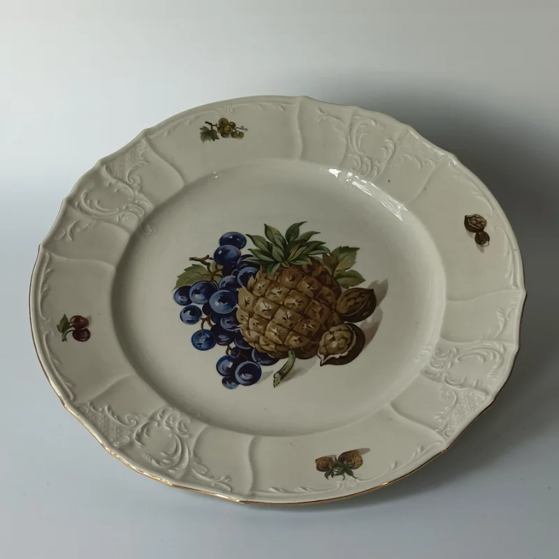 Duży porcelanowy talerz stołowy marki Bernadotte z motywem ananasa i winogron, widok z góry. Detal owocowego wzoru na porcelanowym talerzu: ananas, winogrona i orzechy włoskie na kremowym tle.